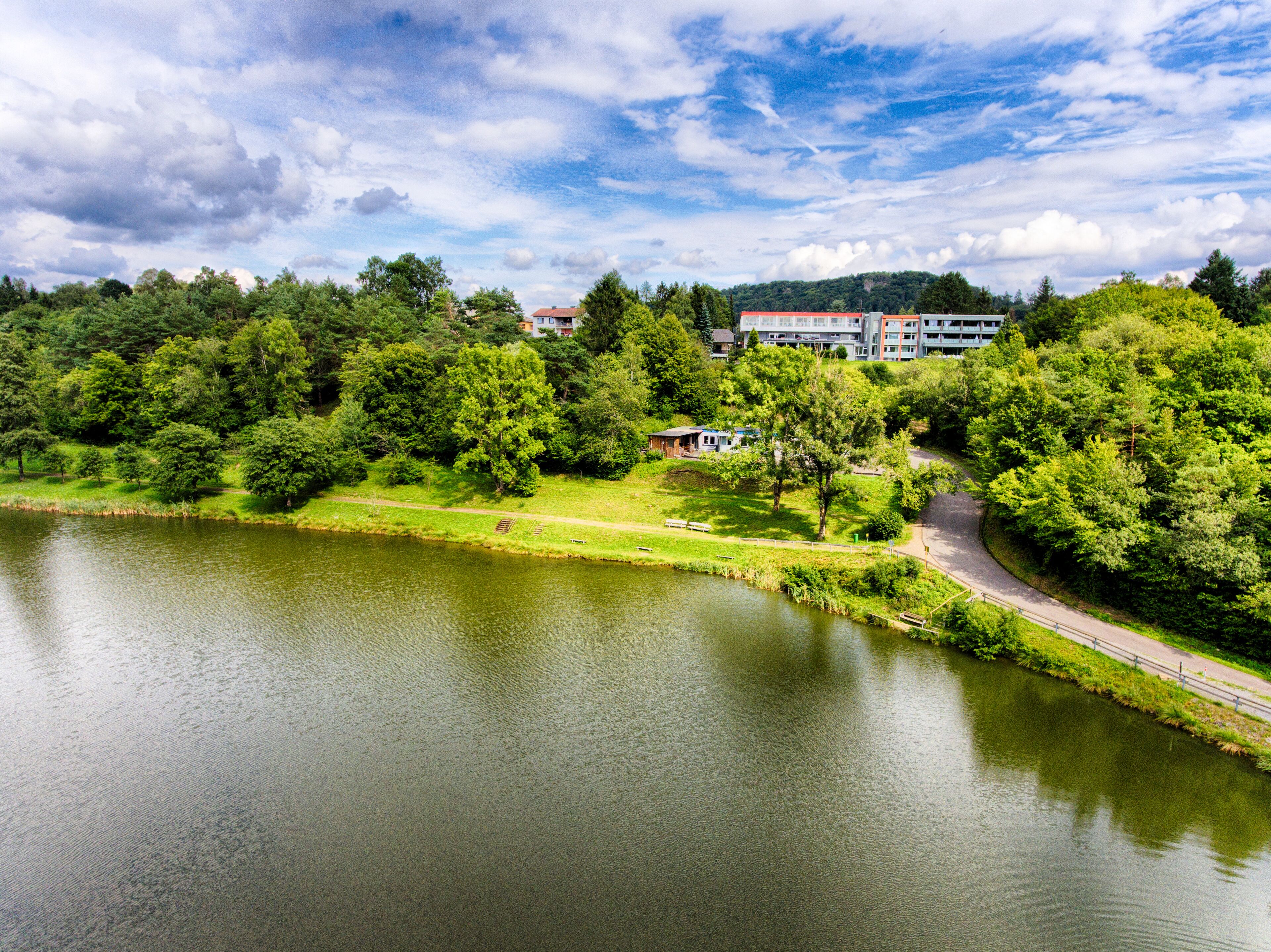 Blick auf einen See mit einem Park, einem Fußweg und einem Gebäude im Hintergrund.