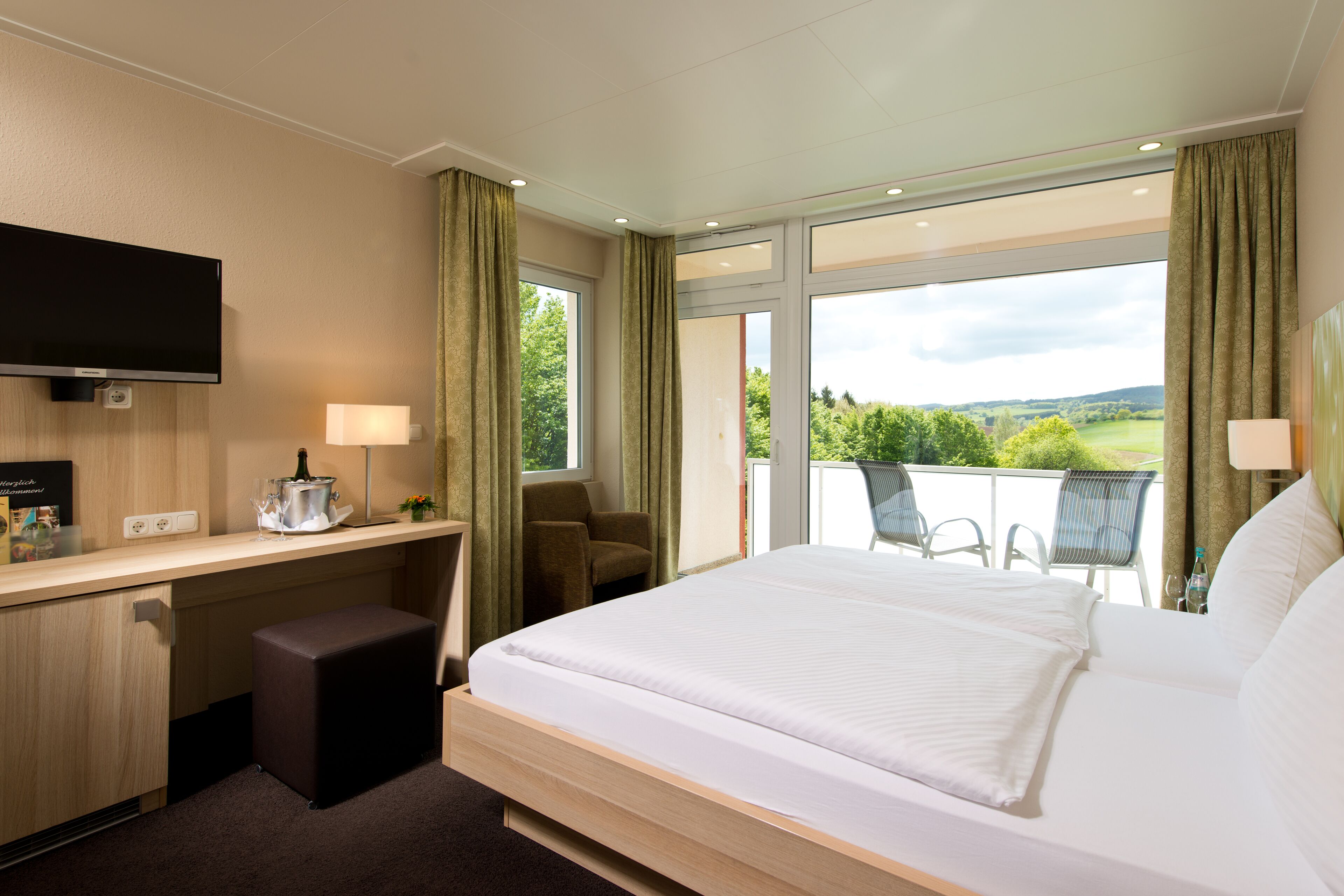 Ein Hotelzimmer mit Bett, Schreibtisch, Fernseher und einem Balkon mit Blick auf die Landschaft.