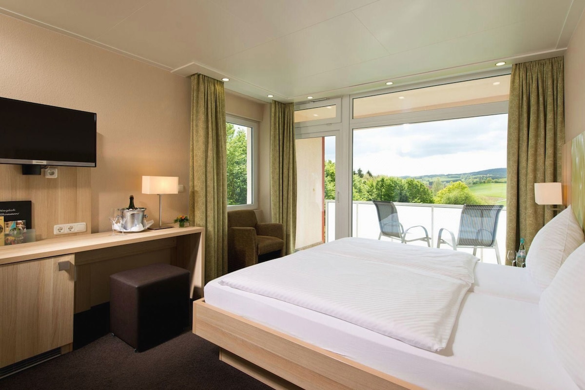 A hotel room with a bed, desk, chair, and a balcony with a view.