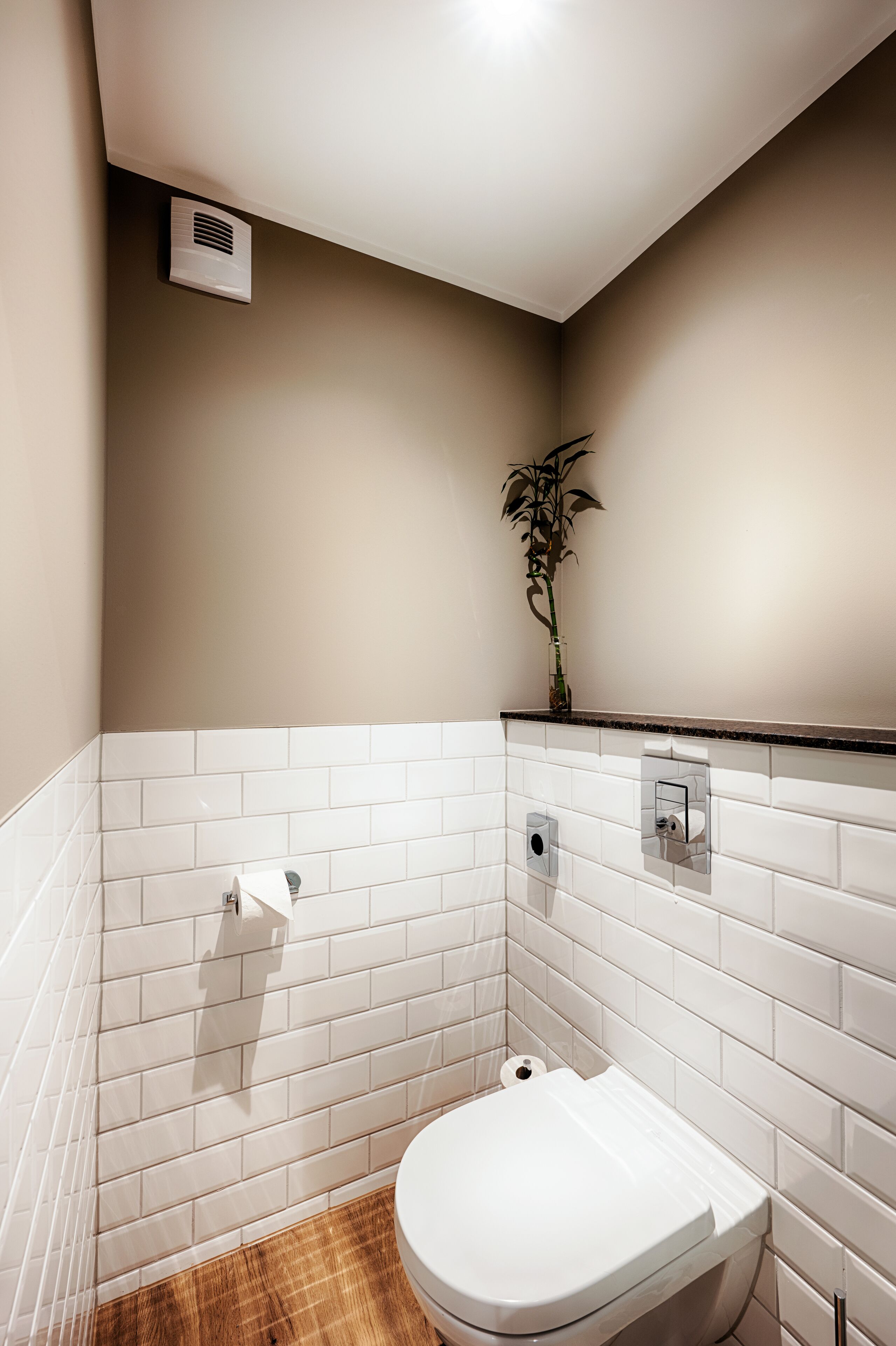 A small, modern bathroom with white tiles, a toilet, a towel dispenser, and a plant.