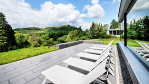 A rooftop terrace with white lounge chairs, a stone wall, and a view of a green landscape and hills. - Seehotel am Stausee (Gerolstein)