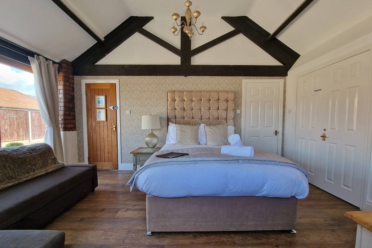 A bedroom with a large bed, a nightstand, a lamp, a wooden bench, and a door.