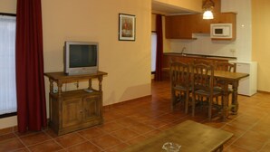Ein kompakter Wohnraum mit Fernseher auf einem Holzständer, einem Esstisch mit Stühlen, einer Küchenzeile mit Mikrowelle und einem roten Vorhang.