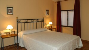 Ein ordentlich bezogenes Bett in einem Zimmer mit zwei Nachttischen, jeweils mit einer Lampe, einem Fenster mit Vorhängen und zwei gerahmten Bildern an der Wand.