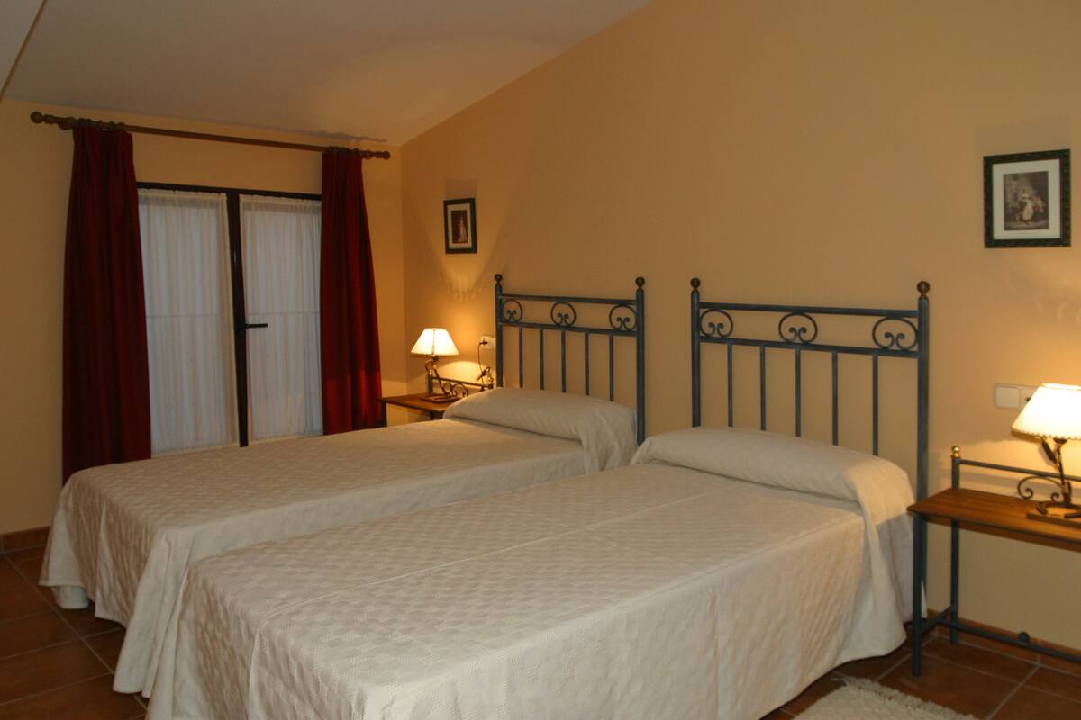 A bedroom with two single beds, a nightstand with a lamp, a window with curtains, and a picture on the wall.