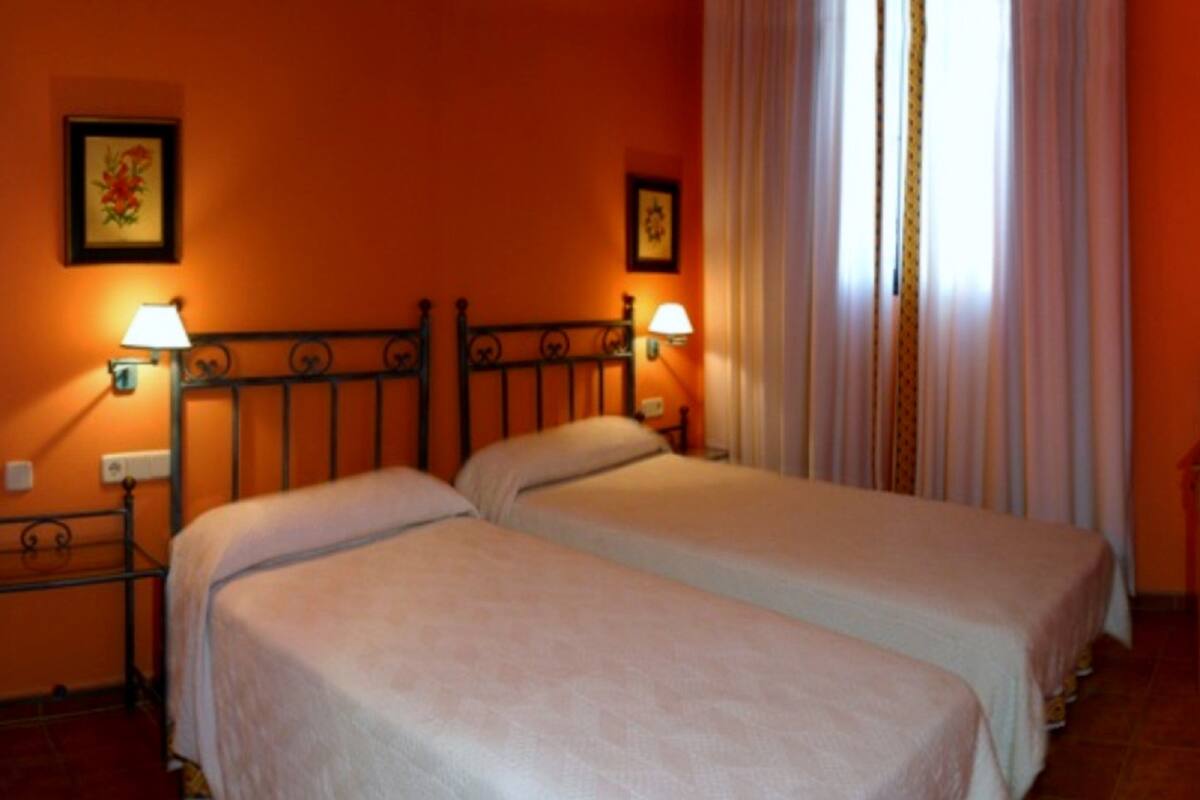 A room with two beds, each with a headboard and bedside lamps. There are two framed pictures on the wall, and a window with curtains.