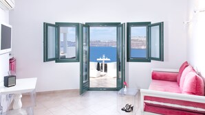 Una habitación con un sofá blanco, un sofá rosa, una mesa con una radio y vistas al mar a través de ventanas abiertas.