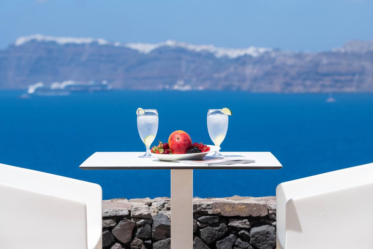 A table with two glasses of a clear beverage, a plate of fruit, and a menu on a balcony overlooking a body of water and cliffs.