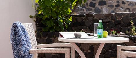 A round table with a stone wall backdrop, two wicker chairs, a blue patterned towel, a green bottle, and a stone on the table.
