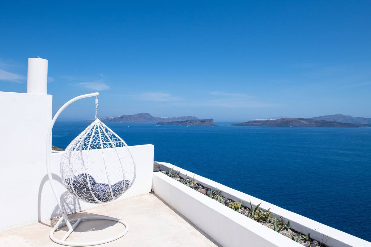 A white outdoor seating area with a spherical hanging chair overlooking a clear blue sea and distant islands.