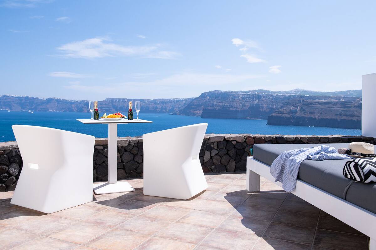A terrace with white furniture, a table with drinks, and a view of the sea and cliffs.
