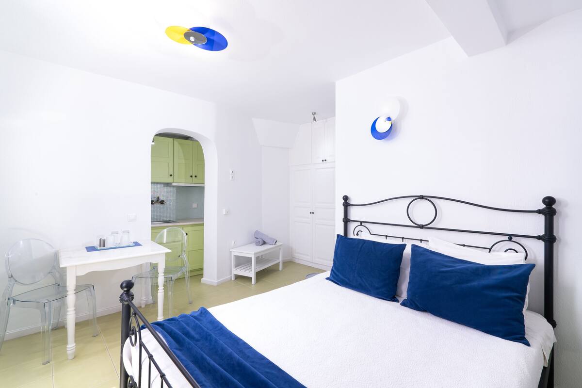 A bedroom with a bed, a desk, and a chair. There is a kitchenette in the background.