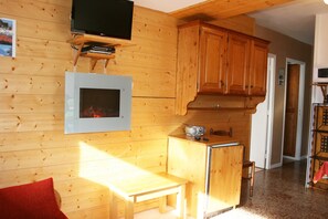 A wooden interior with a television mounted on a shelf, a small table, and a kitchenette area. - Hôtel Le Catinat Fleuri (Guillestre)