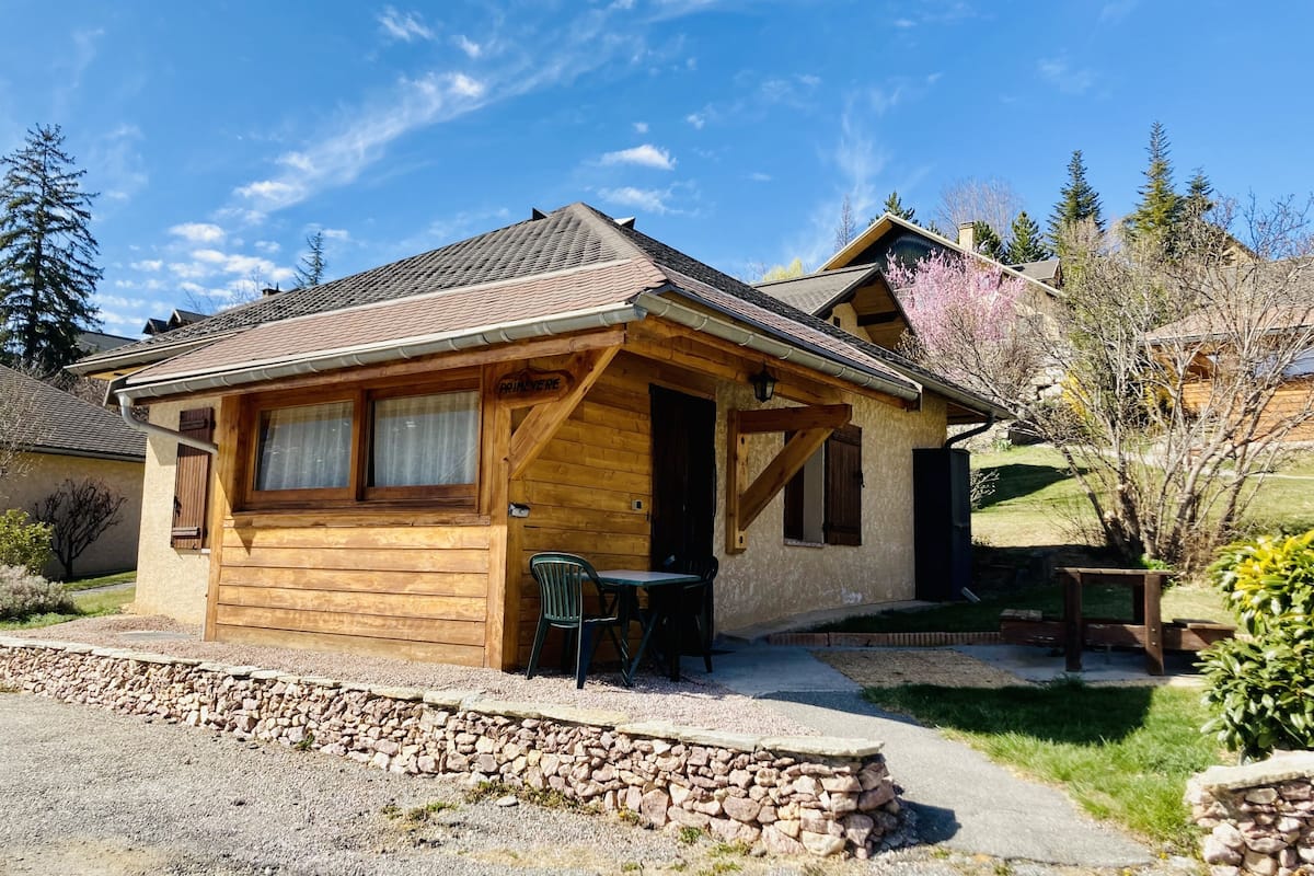 Une cabane en bois avec des fondations en pierre, une véranda et une petite table avec des chaises placée à l’extérieur.