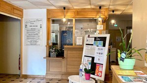 Un espace d’accueil d’hôtel avec un panneau d’affichage présentant les tarifs des chambres, une petite table avec une plante et un sol carrelé.