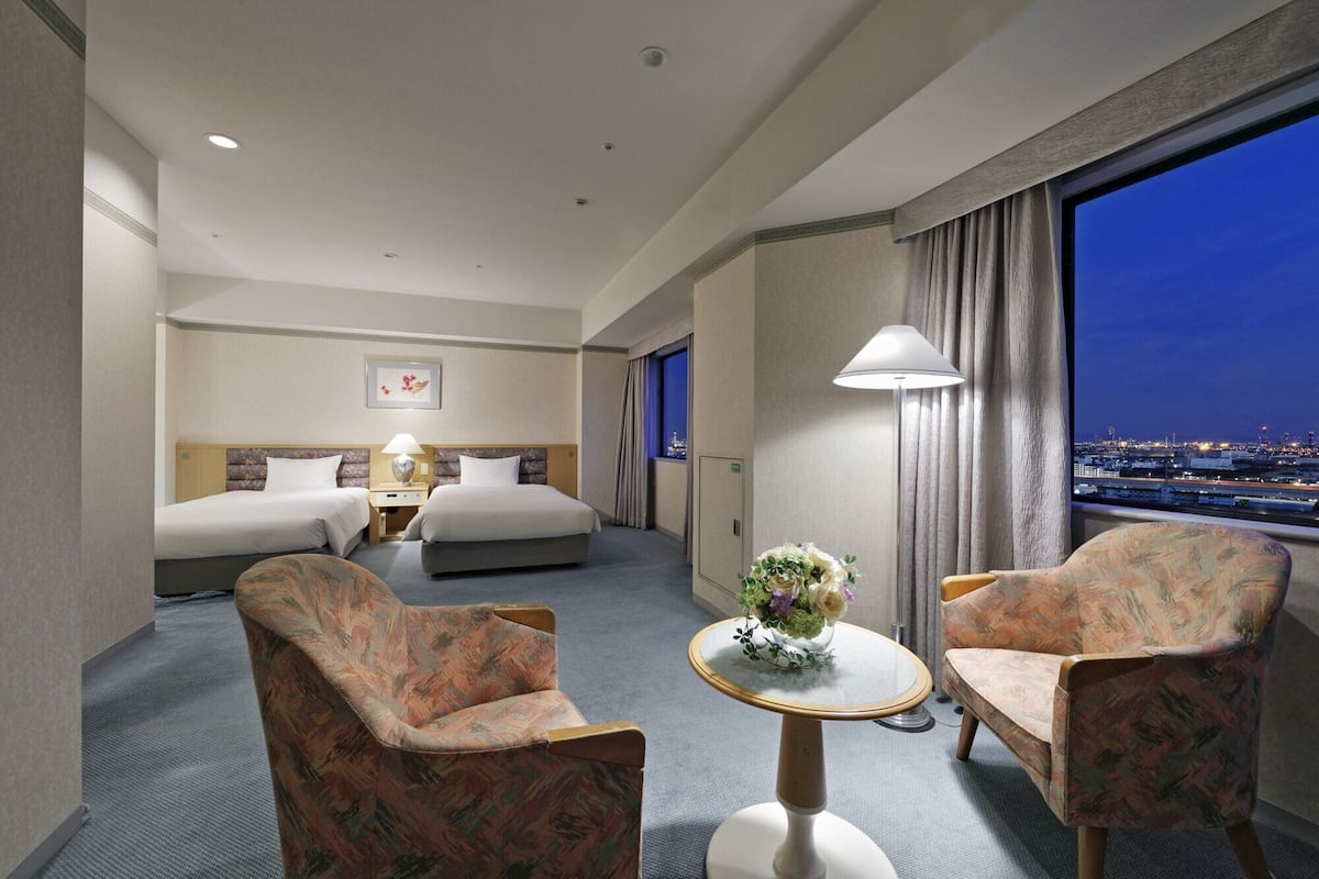 Una habitación de hotel con una cama, dos sillones, una mesita con un jarrón y vistas a la ciudad de noche.
