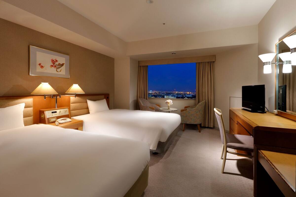 Habitación de hotel con dos camas, un escritorio, una silla y un gran ventanal con vistas a la ciudad.