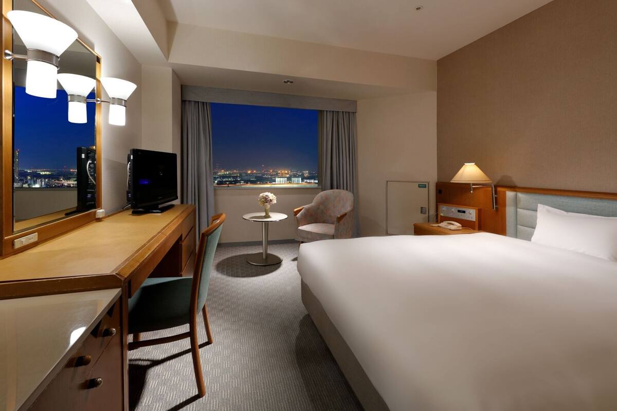 Habitación de hotel con cama, escritorio, silla, televisor y vistas a la ciudad de noche.