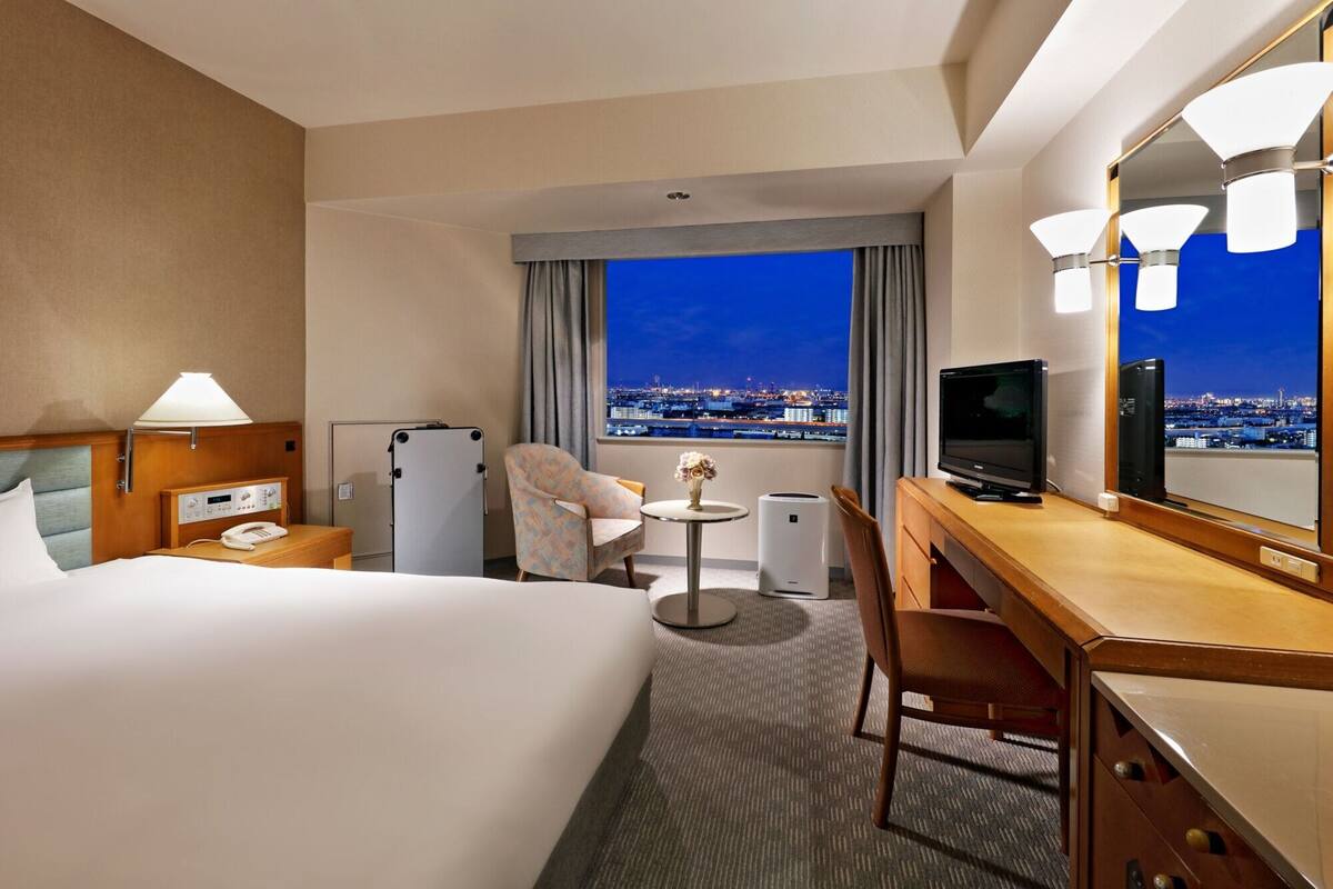 Habitación de hotel con una cama, un escritorio con televisor, una silla, una mesita con un florero y vista a un paisaje urbano nocturno.