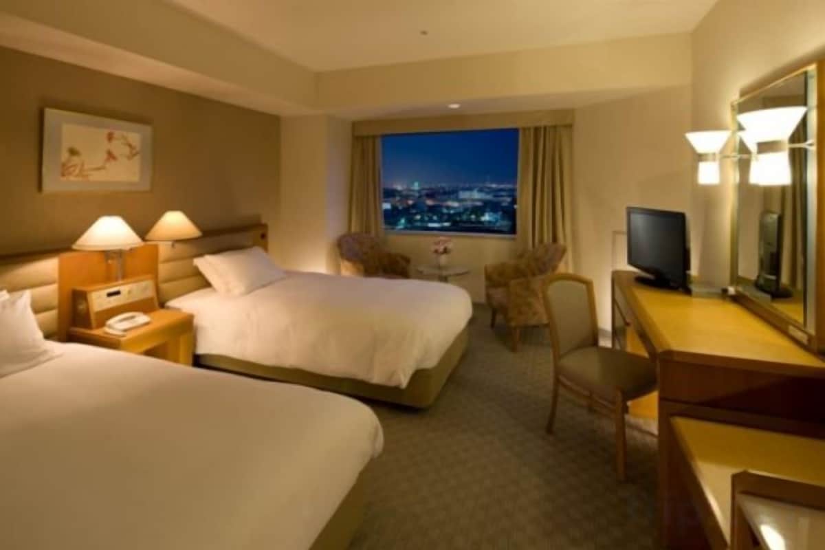 Een hotelkamer met twee bedden, een bureau, een stoel, een televisie en uitzicht op de stad 's nachts.