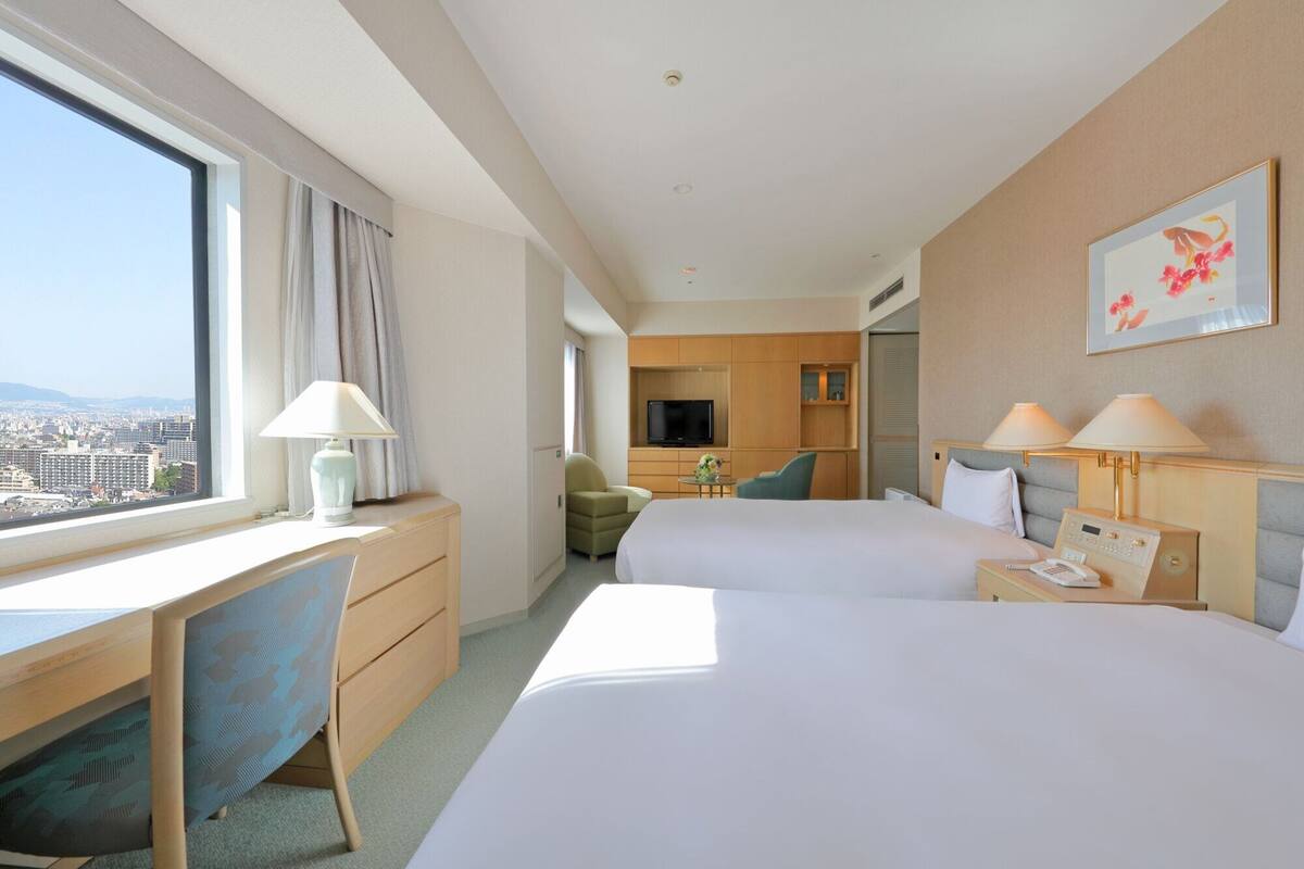 Habitación de hotel con dos camas, un escritorio, una silla, un televisor y vista a la ciudad.