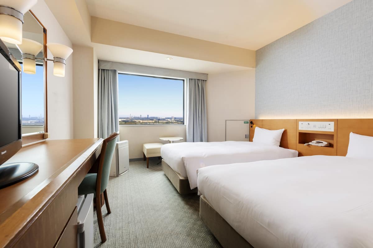 Habitación de hotel con dos camas, un escritorio con silla, una mesita y un espejo, con vista a la ciudad a través de la ventana.