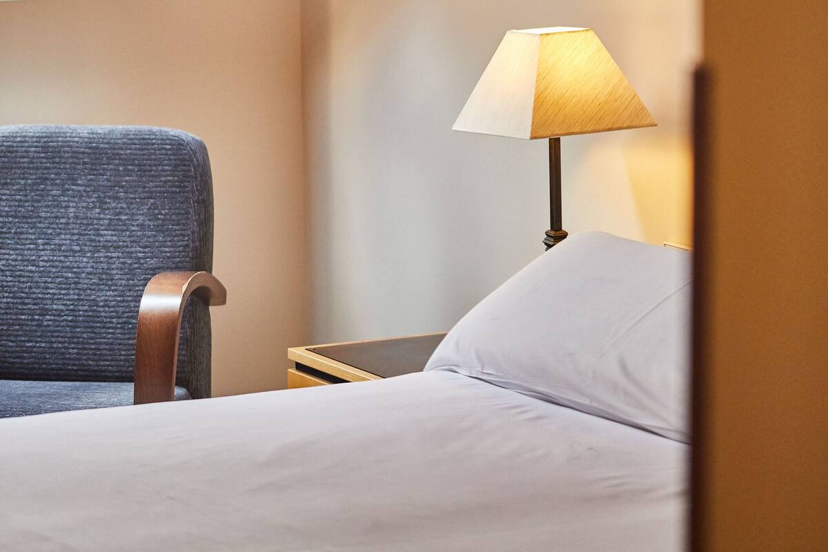 A room with a sloped ceiling, a bed, a chair, and a nightstand with a lamp.