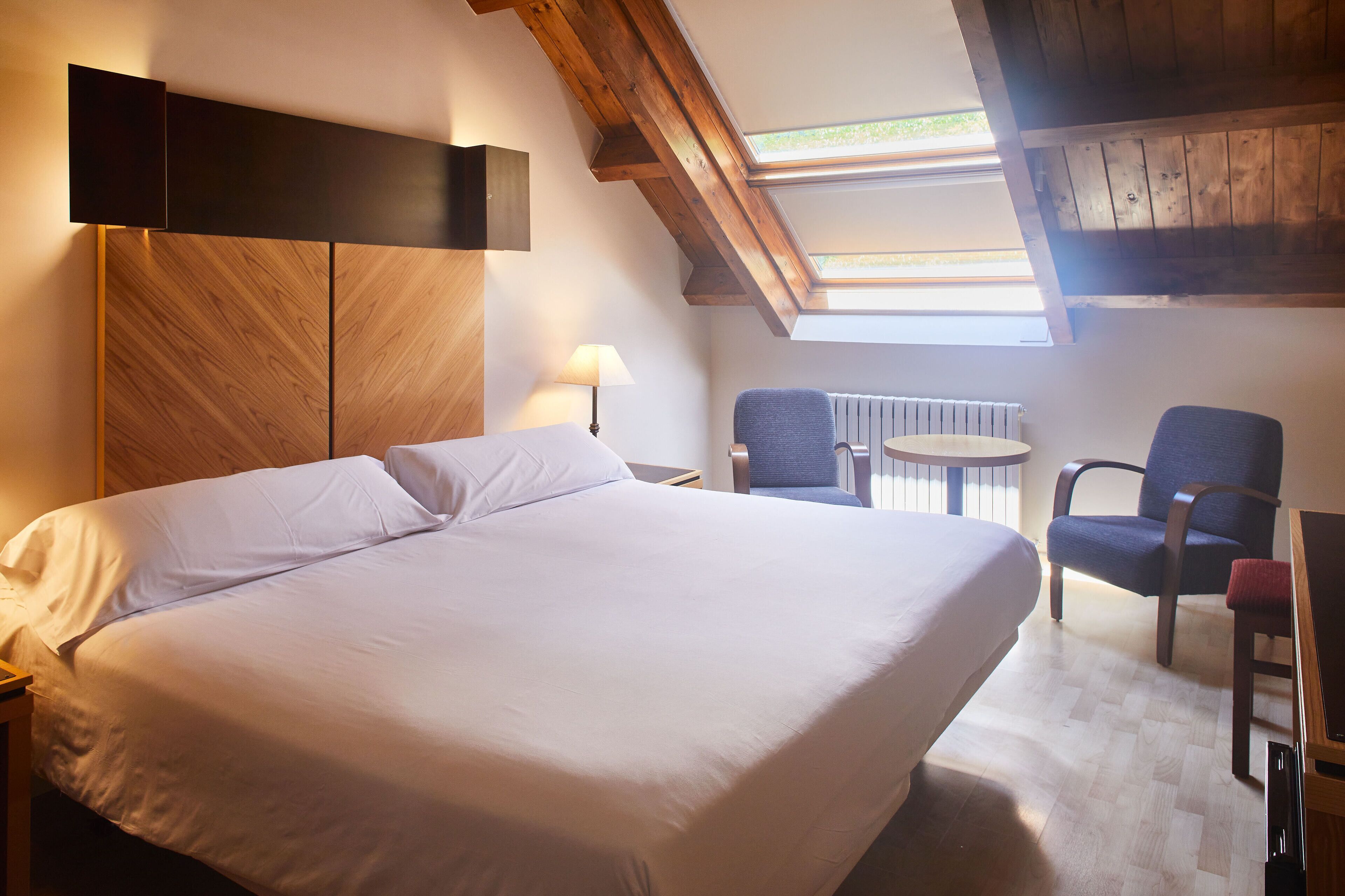 Habitació d'hotel amb un llit gran, una taula petita amb cadires, un fanal i bigues de fusta.