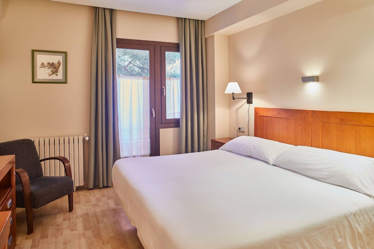A hotel room with a bed, a chair, a desk, a lamp, and a window with curtains.