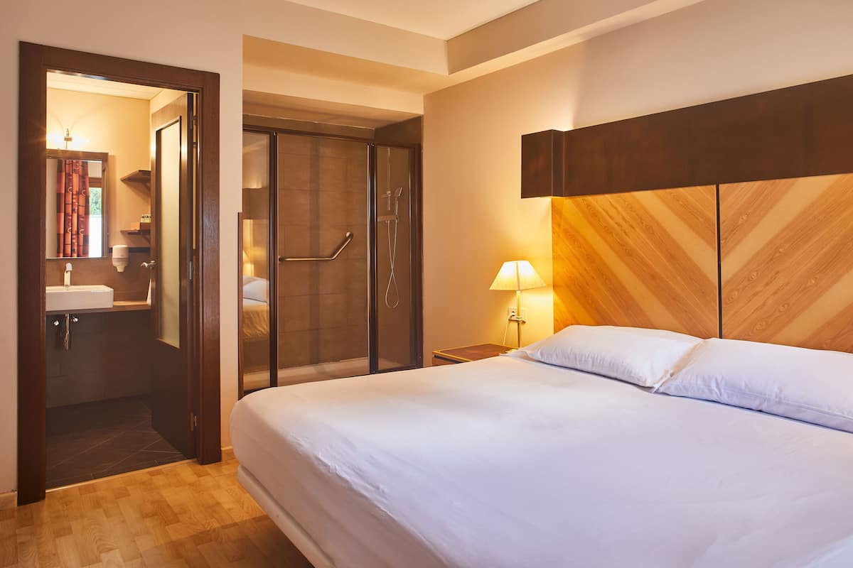 A hotel room with a bed, a nightstand with a lamp, a wooden headboard, and a shower area.