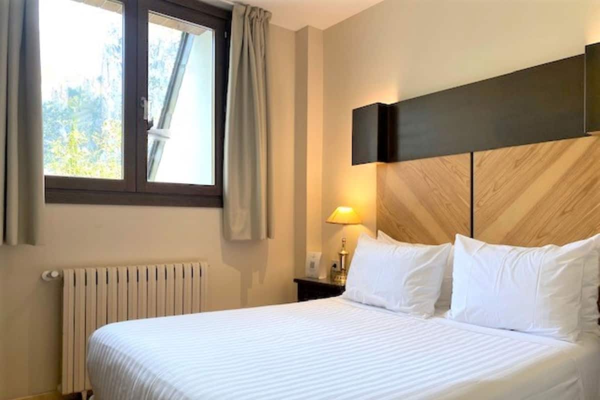A hotel room with a large bed, a window with curtains, a bedside lamp, and a radiator.