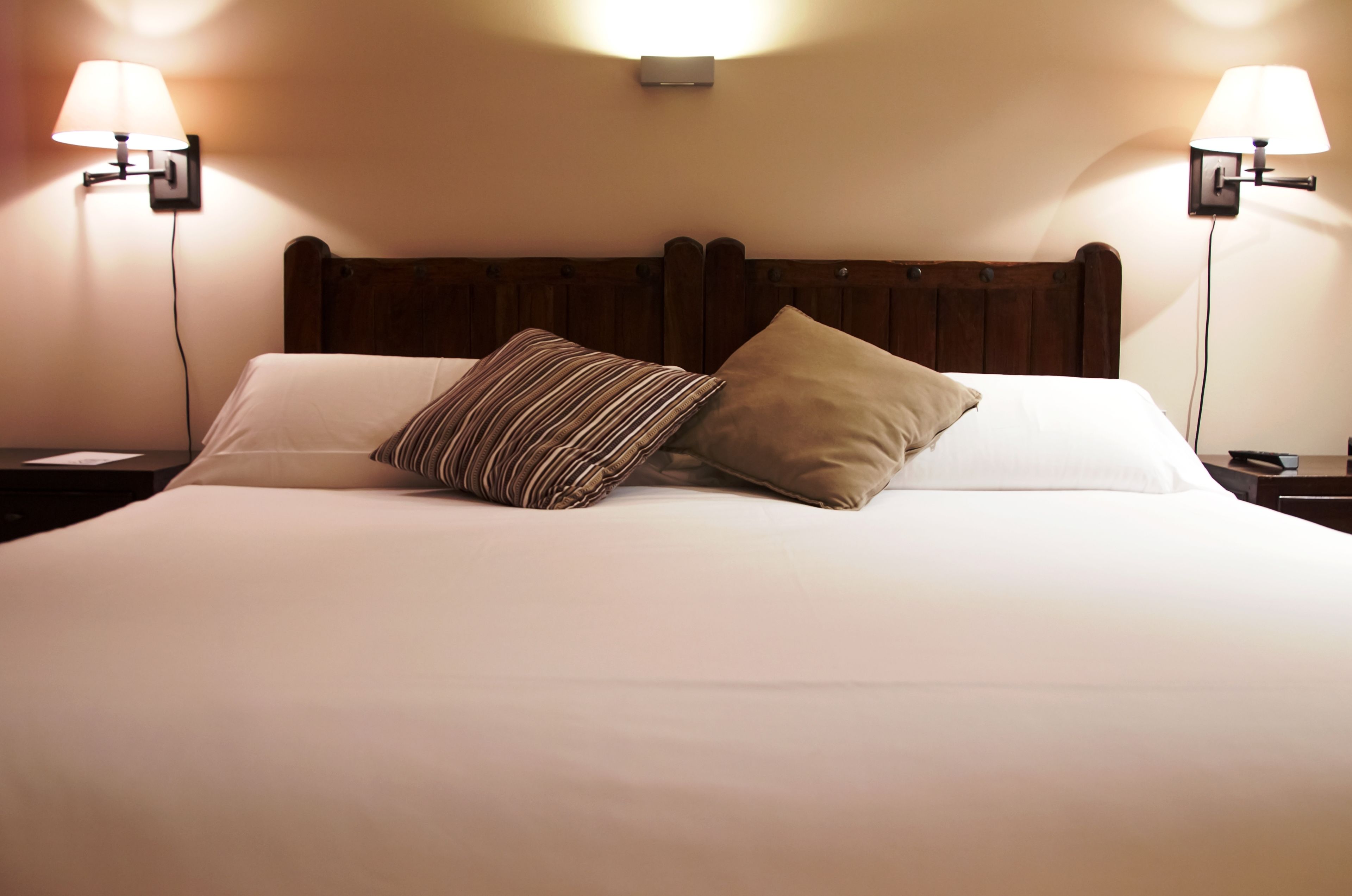 Una cama bien hecha con cabecera de madera, dos lámparas de noche y un aire acondicionado de pared.