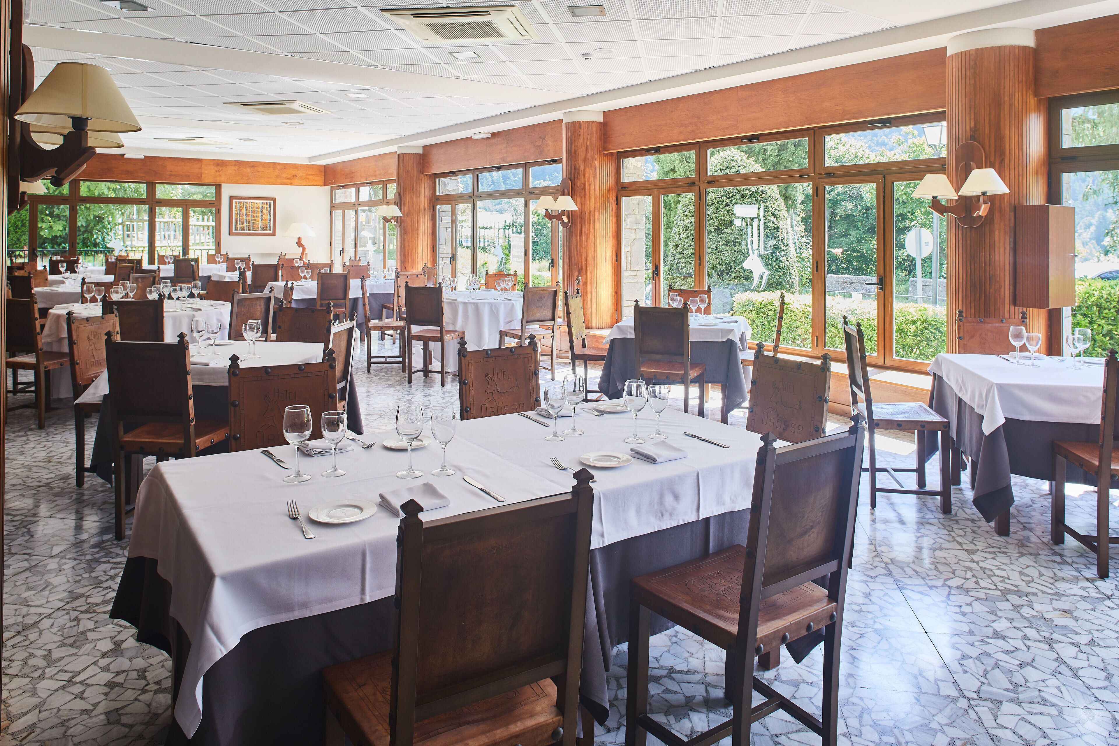 Un comedor espacioso con varias mesas dispuestas para comer, ventanas amplias que ofrecen vistas a la vegetación y paneles de madera en las paredes.