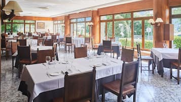 Un comedor espacioso con varias mesas dispuestas para comer, ventanas amplias que ofrecen vistas a la vegetación y paneles de madera en las paredes.