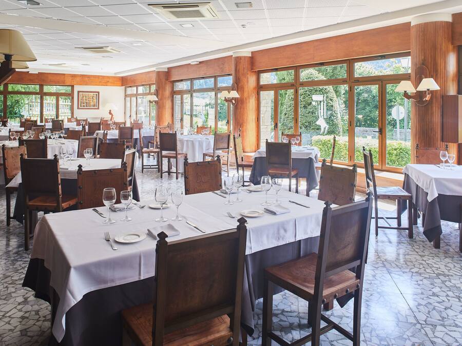 Un comedor espacioso con varias mesas dispuestas para comer, ventanas amplias que ofrecen vistas a la vegetación y paneles de madera en las paredes.