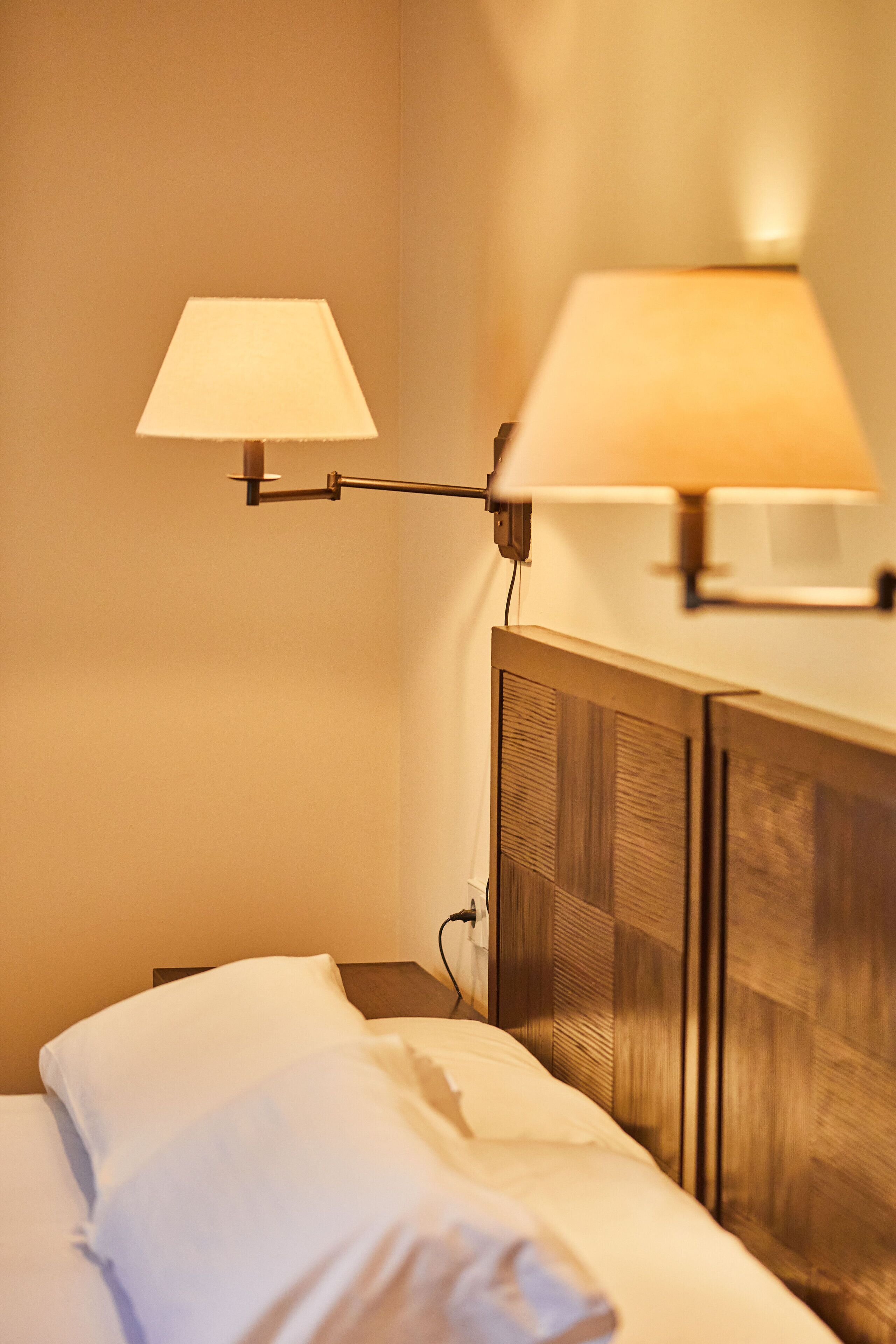 Una cama bien hecha con sábanas blancas, una mesita de noche de madera y dos lámparas de pared que ofrecen una luz tenue.
