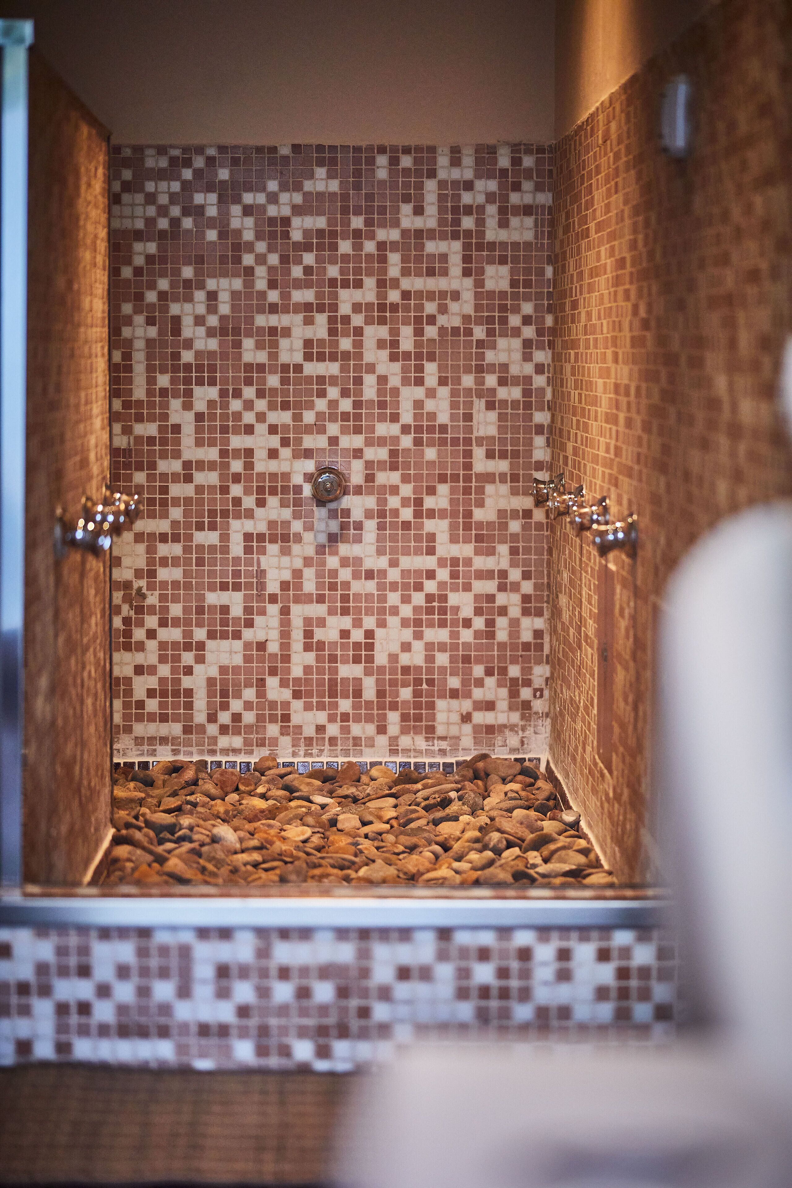 Una ducha con azulejos y un diseño de mosaico, un grifo metálico y un suelo revestido de piedras.