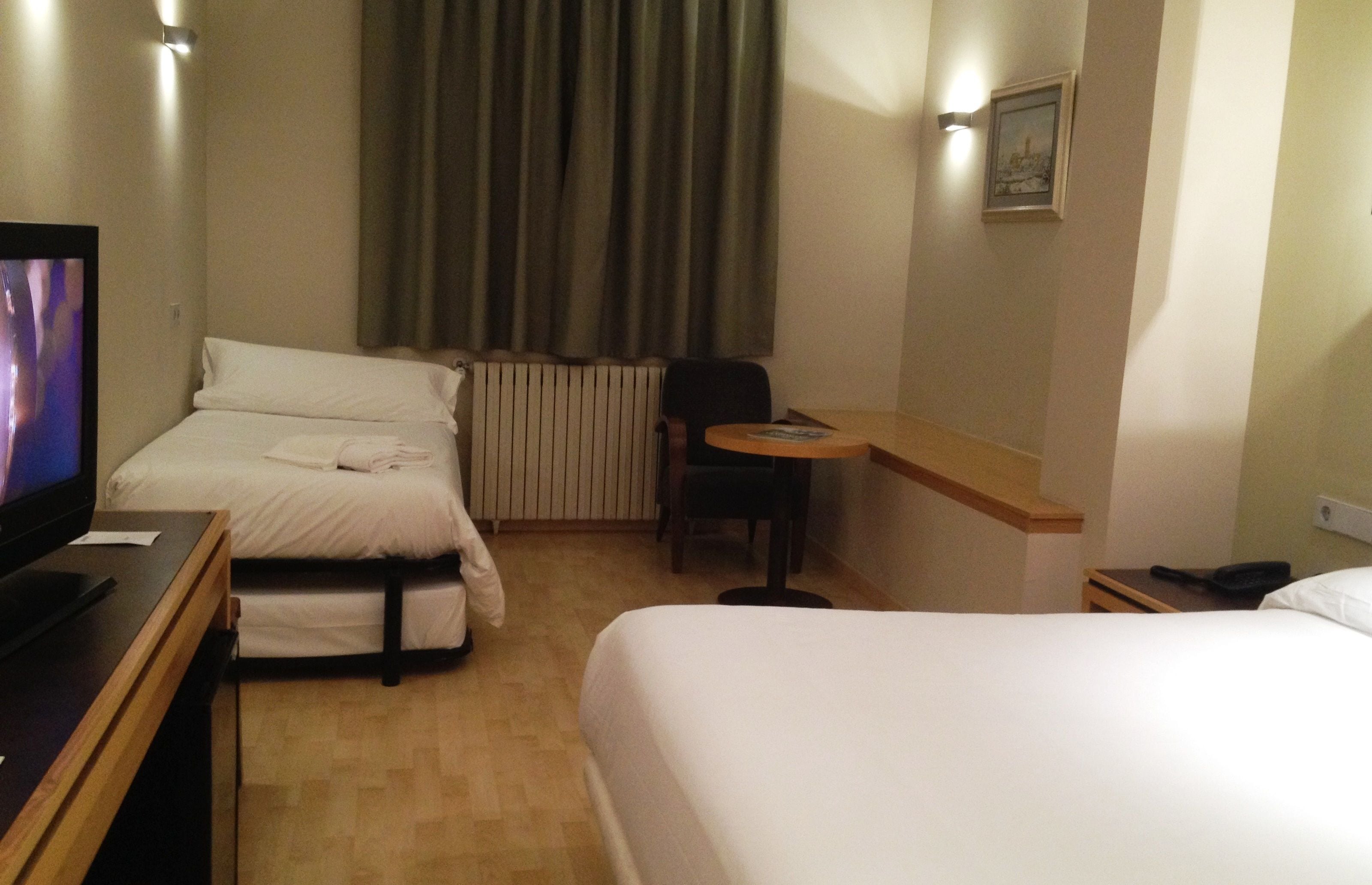 Habitación de hotel con dos camas, un escritorio, una silla, televisión y ventana con cortinas.