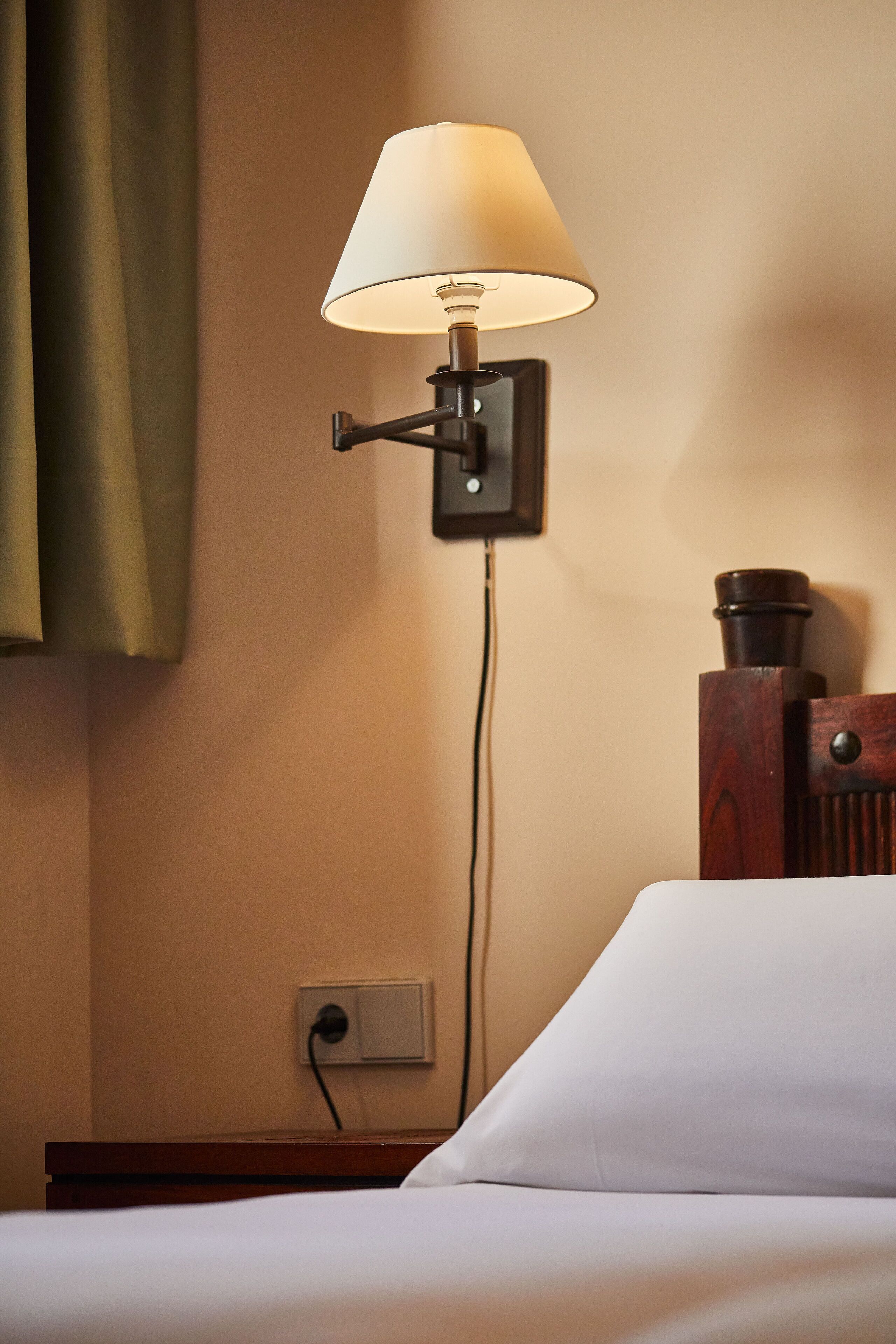 A bed with a white sheet, a wall-mounted lamp, and a power outlet.
