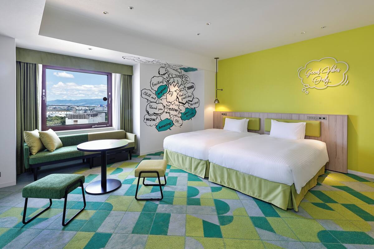 Habitación de hotel con una cama grande, un sofá, una mesita y un ventanal con vistas a edificios.