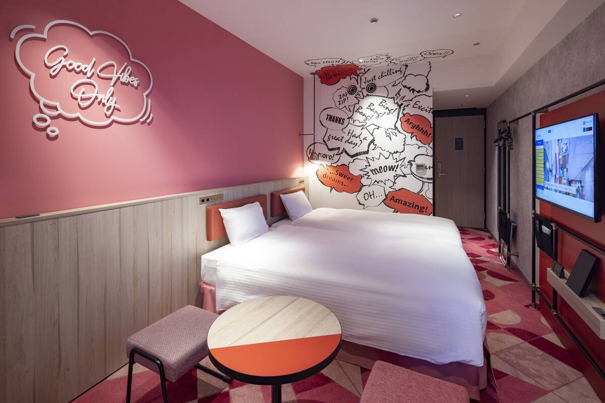 Habitación de hotel con una cama grande, una mesita redonda pequeña, una pared rosa con un letrero que dice 'Good Vibes Only', y una pared con varias palabras y frases.