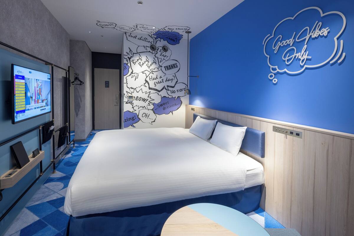 Una habitación de hotel moderna con una cama grande, un televisor a la izquierda y una pared con frases motivacionales.