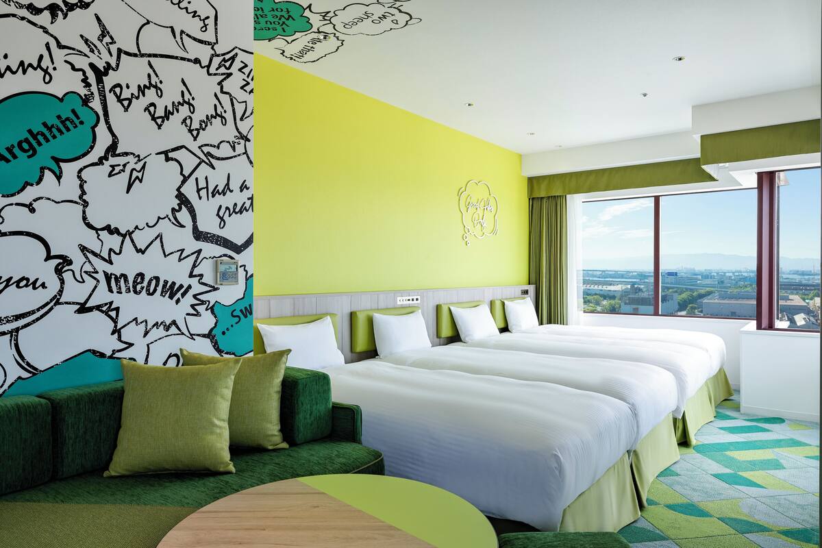 Habitación de hotel con dos camas, un sofá verde, un ventanal grande con vista a la ciudad y una pared decorada con bocadillos de diálogo.