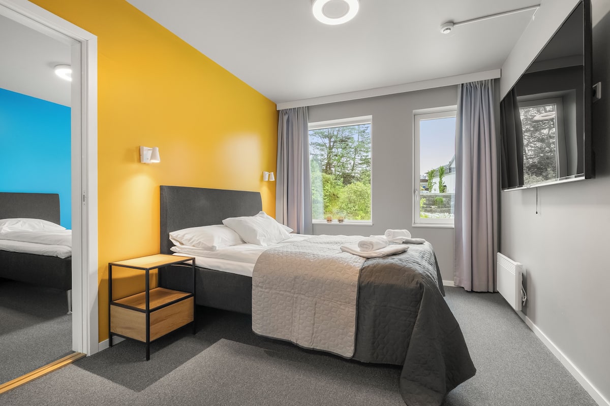A modern bedroom with a bed, a bedside table, a window with curtains, and a wall-mounted TV.
