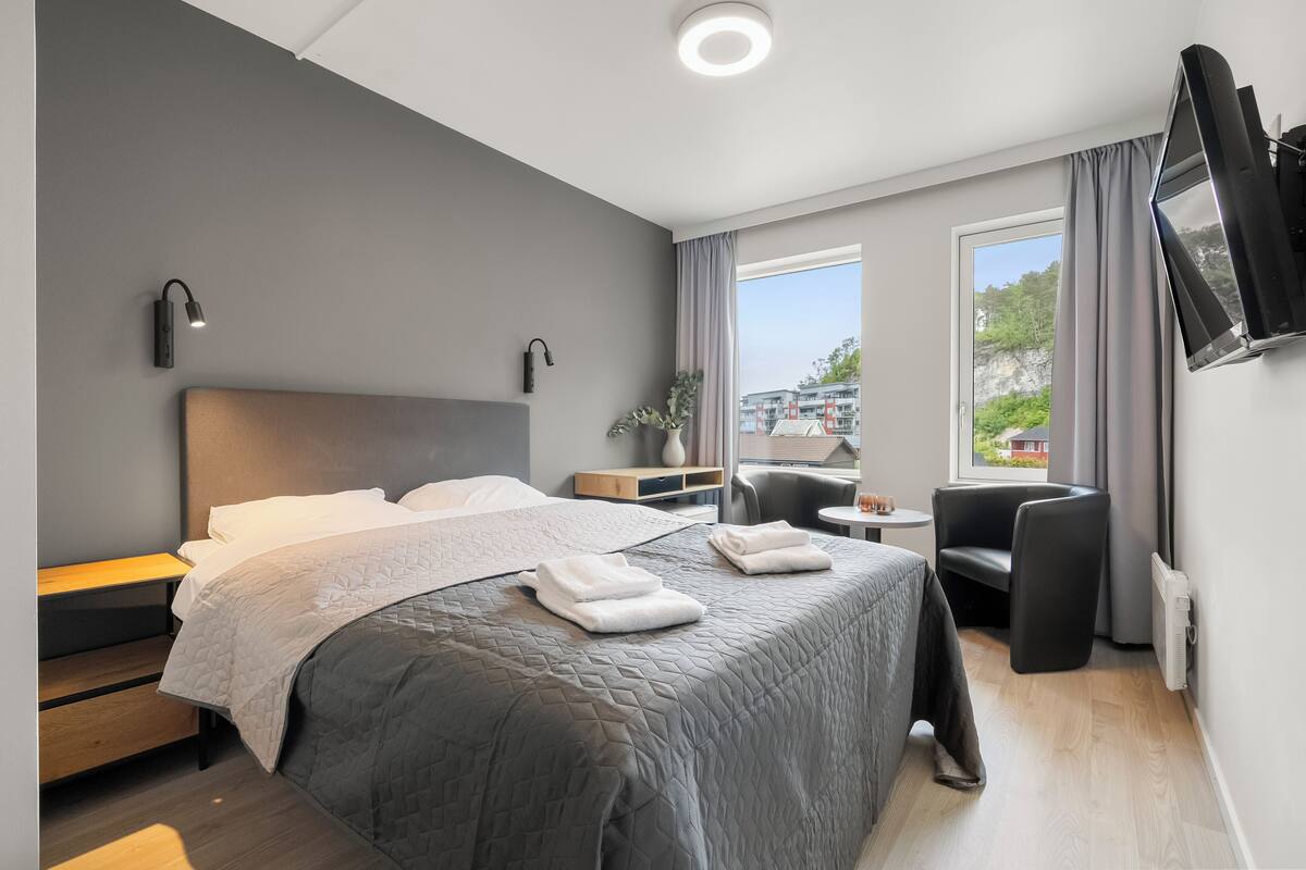 A modern hotel room with a large bed, bedside tables, a small table, a chair, and a TV mounted on the wall.