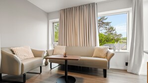 A modern living room with a beige sofa, a small round table, and a view of greenery through large windows. - Kinn Hotell Florø (Kinn)