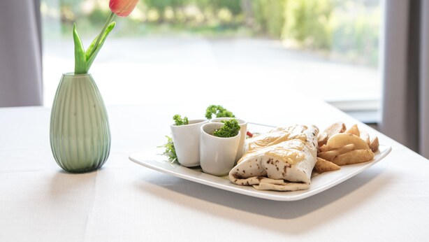 A plate of food with a rolled item, two small white cups, and a green vase with a single flower. - Kinn Hotell Florø (Kinn)