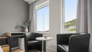 A modern hotel room with a bed, a small desk, two black leather chairs, a round table, and a window with a view of a cliff. - Kinn Hotell Florø (Kinn)