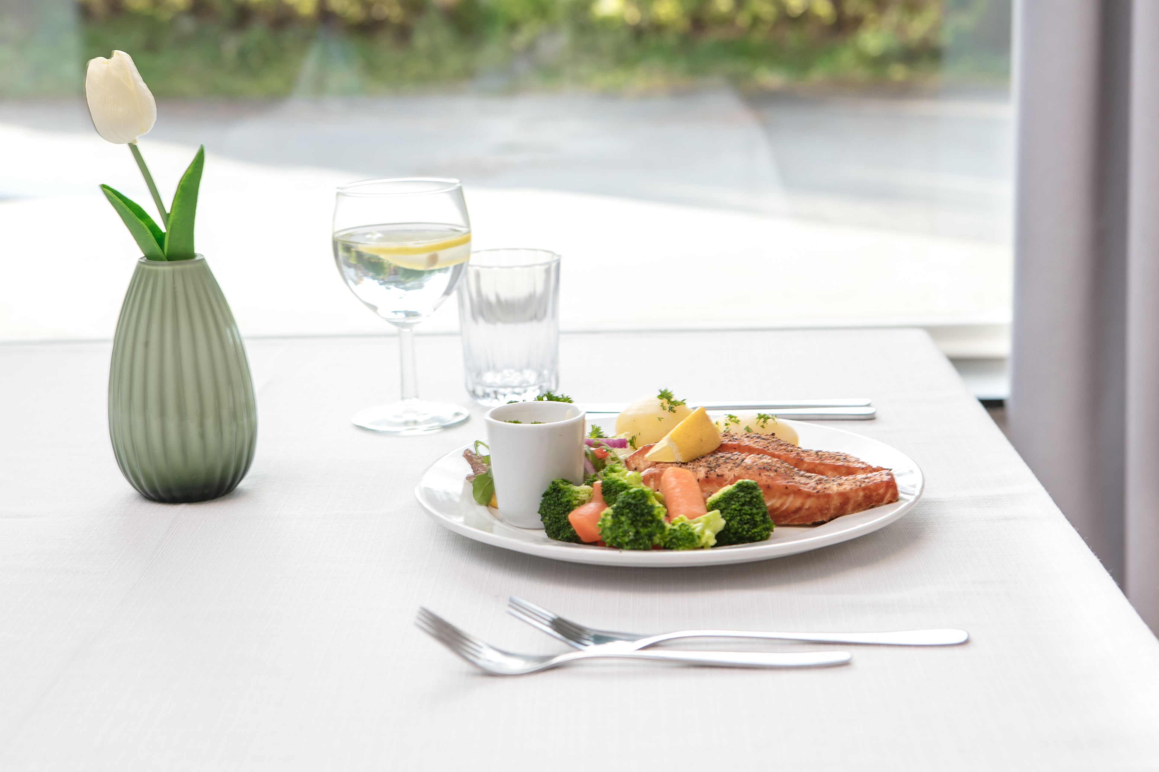 A dining table with a plate of food, a glass of water, a small vase with a flower, and a fork.
