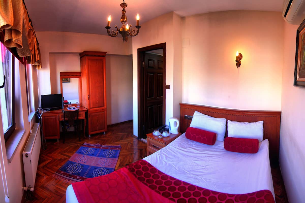 A hotel room with a bed, a desk with a computer, a chair, a window with curtains, a radiator, a wall-mounted light, and a painting.